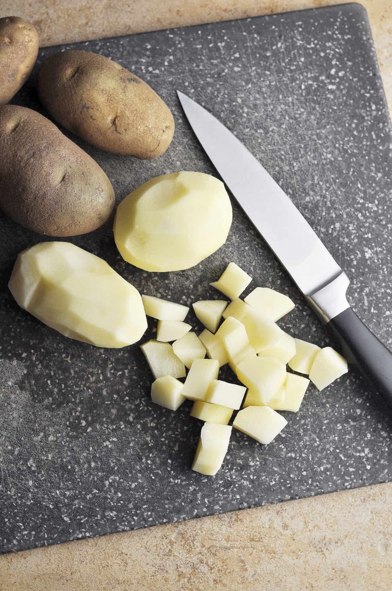 Raw potatoes with skin on, peeled, and cut into chunks on cutting board with sharp knife.