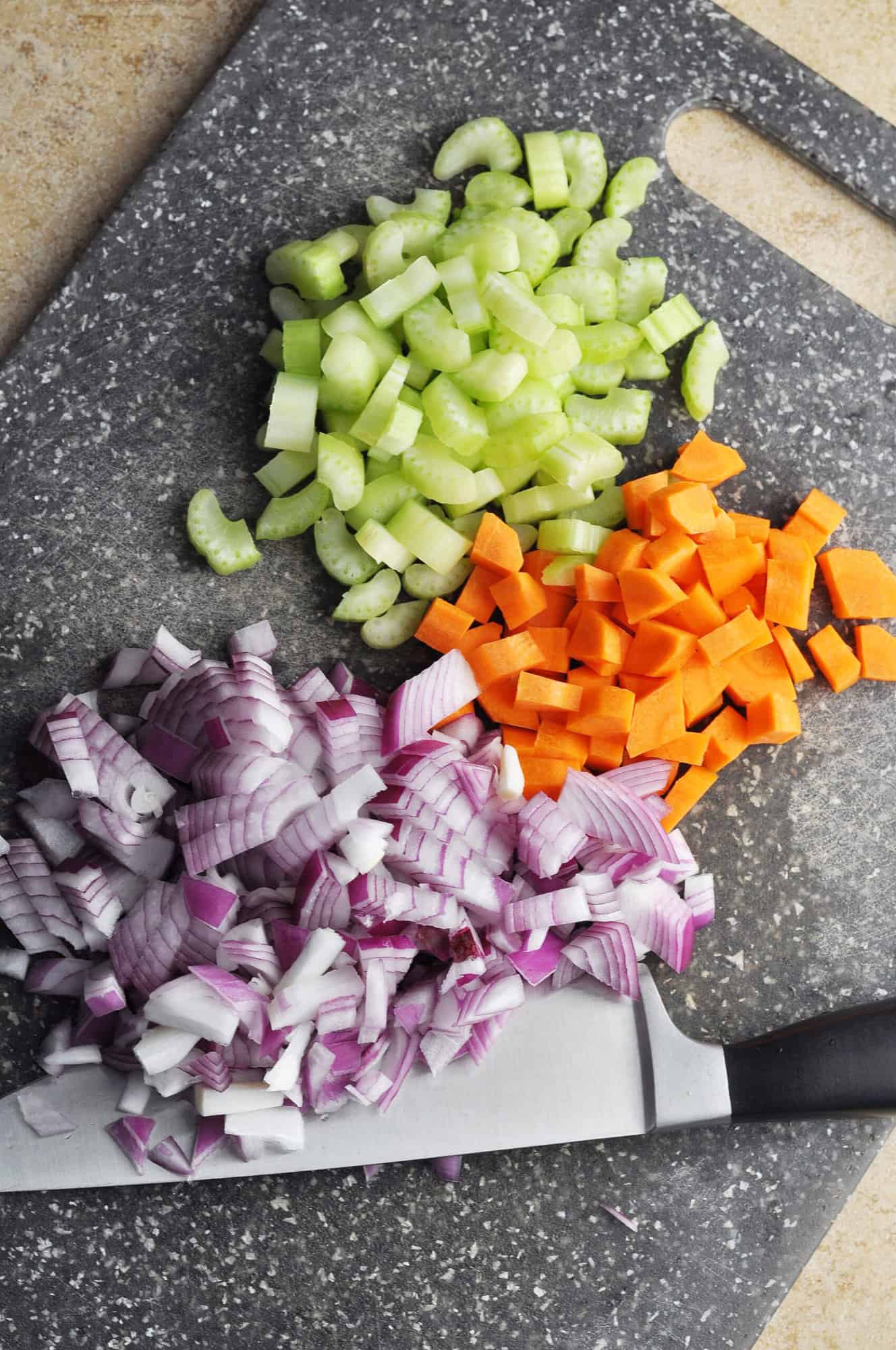 Chopped onion, carrot, and celery on cutting board with knife.