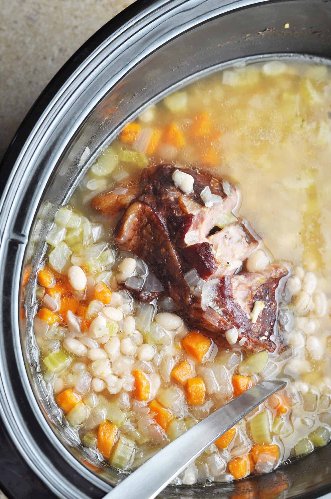 Cooked navy bean soup with carrots and celery and ham hock in slow cooker.