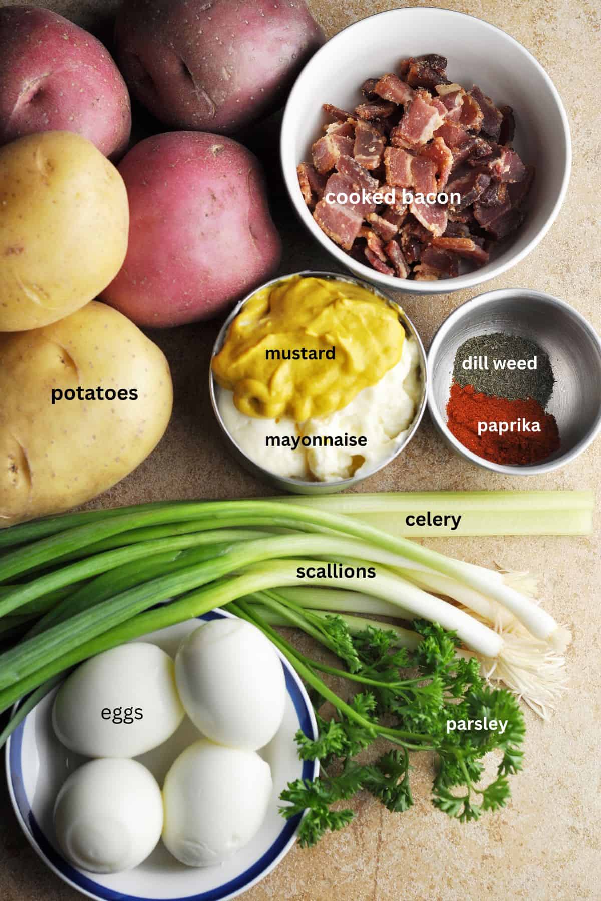 Ingredients for potato salad, including boiled eggs, potatoes, bacon, parsley, celery, scallions, dry dill, paprika, mustard, and mayonnaise.